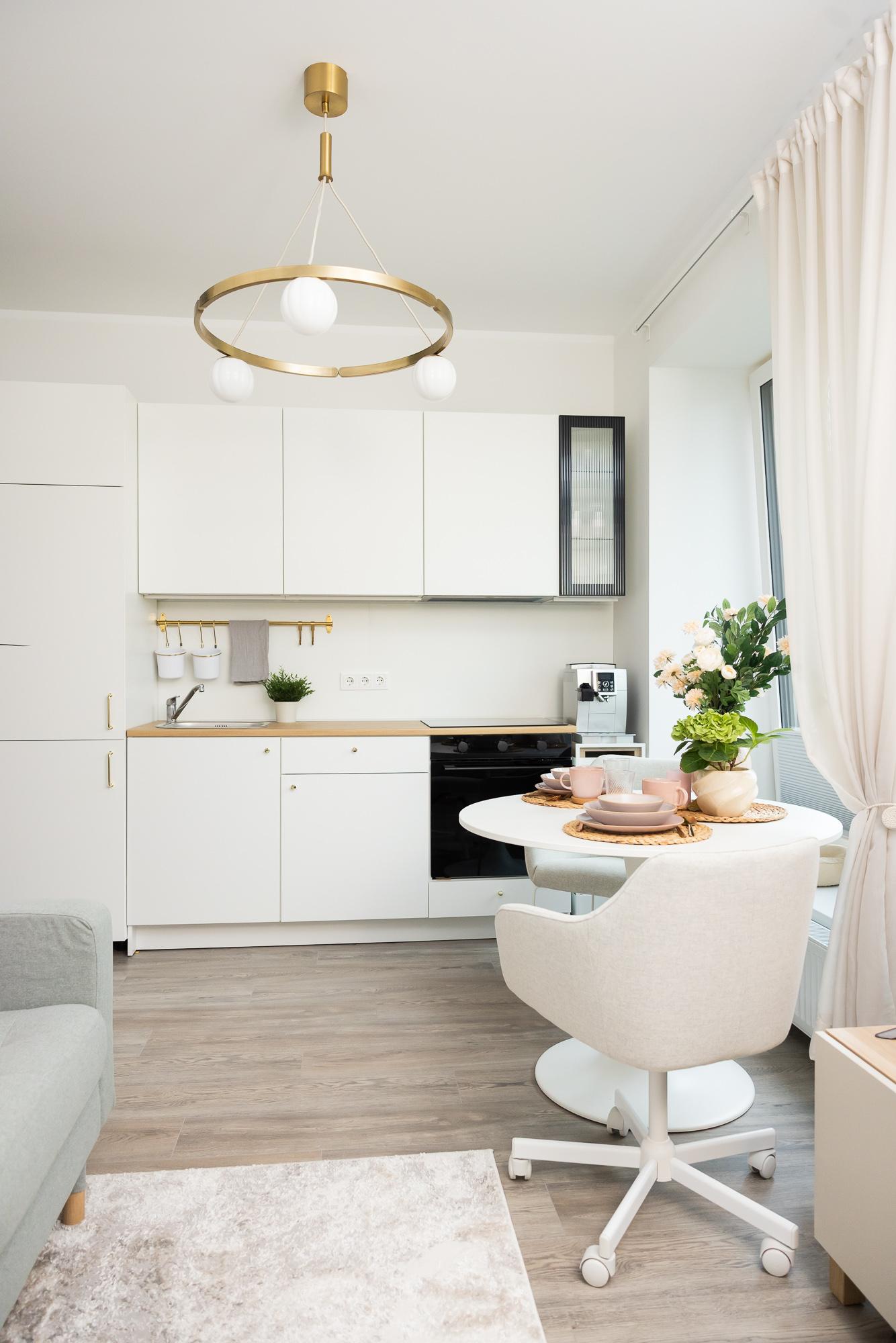 Bright and compact open-plan kitchen with white cabinetry, a small dining area, soft textiles and contemporary lighting, creating a cosy and functional living space.