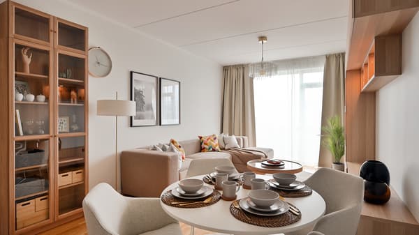 Bright and contemporary open-plan living and dining area with warm wooden finishes, built-in shelving, a round dining table set for four, soft neutral furnishings, and large windows allowing natural light into the modern apartment interior.
