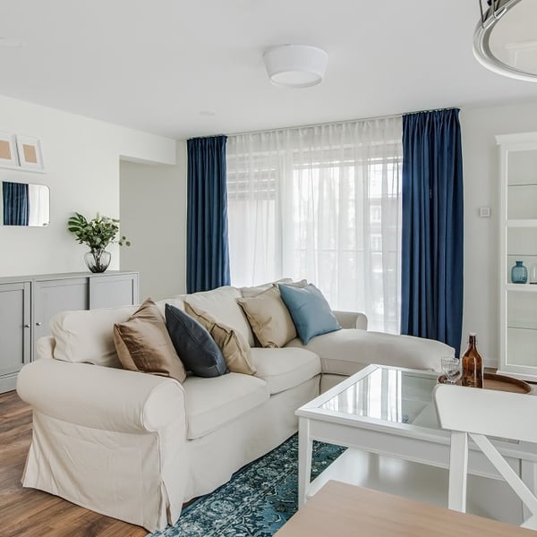 Bright and elegant living room featuring a large off-white sectional sofa adorned with assorted throw pillows in shades of brown, navy, and blue. The sofa sits on a teal patterned area rug, complemented by sheer white curtains and deep navy drapes covering a large window. A white coffee table with a glass top stands in front of the sofa. In the background, a gray sideboard with a vase of greenery and wall-mounted mirrors adds a decorative touch. The space has a cozy, modern-classic style with a soft and inviting atmosphere.