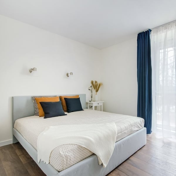 Bright and minimalist bedroom featuring a light gray upholstered bed with a beige bedspread and a soft white throw blanket at the foot. The bed is decorated with four pillows in black, mustard yellow, and patterned designs. On the right, a small white nightstand holds a silver lamp and a vase with dried pampas grass. Wall-mounted reading lights are positioned above the bed, and a large window with sheer white curtains and dark navy drapes lets in natural light. The room has a clean, modern aesthetic with warm wood flooring.