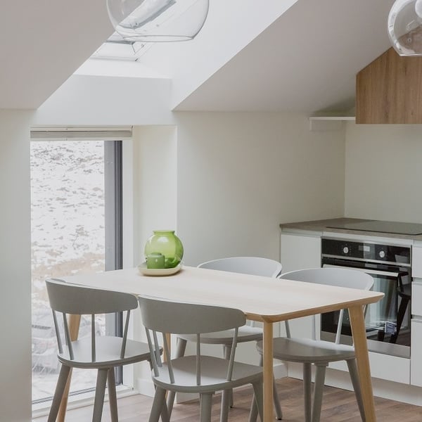 Bright and minimalist kitchen-dining area with a light wood rectangular table surrounded by four pale gray chairs. A green glass vase sits in the center of the table, adding a fresh accent. The room features a slanted ceiling with a large skylight and a tall window that let in plenty of natural light. In the background, a modern built-in oven and countertop complete the compact, clean-lined kitchen setup. The space has a Scandinavian aesthetic with soft tones and natural materials.