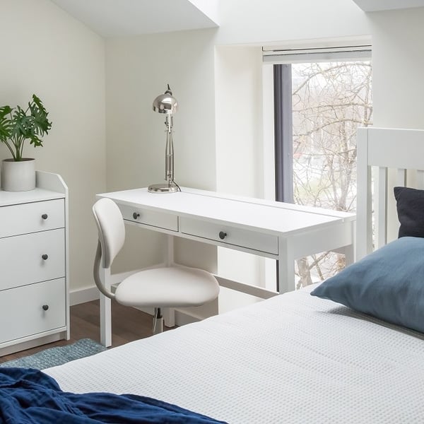 Bright and minimalistic bedroom corner featuring a white desk with black knobs, a modern white swivel chair, and a chrome desk lamp. A tall window next to the desk lets in natural light and shows bare tree branches outside. To the left, there is a small white chest of drawers with a potted plant on top. Part of a white bed with blue and navy bedding is visible in the foreground. The space feels clean, cozy, and functional.