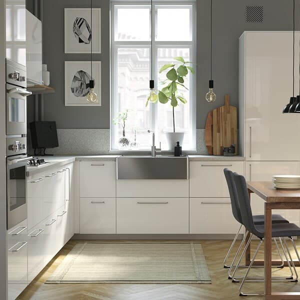 Bright and sleek IKEA kitchen in a U-shaped layout with white cabinets, a farmhouse sink, and a wooden dining area by the window.

