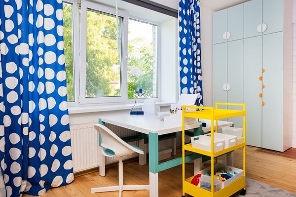 Bright children’s workspace with desk, rolling storage cart, light blue wardrobe, and blue curtains