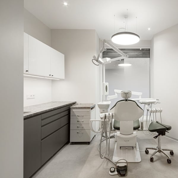 Bright dental treatment room with green dentist chair, overhead lamp, and large frosted window, surrounded by minimalist cabinetry.