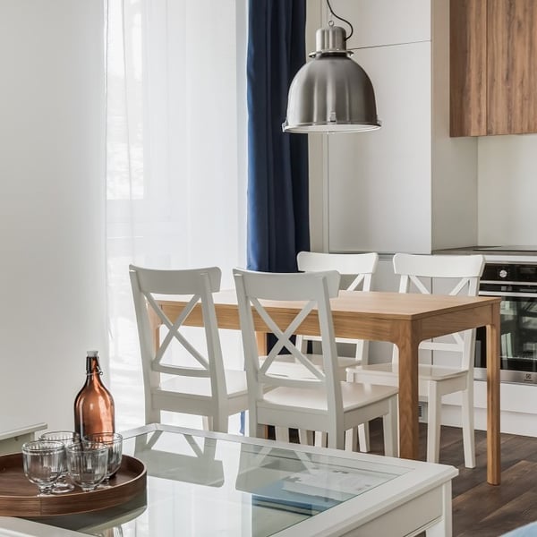 Bright dining and kitchen area featuring a wooden dining table surrounded by four white cross-back chairs. A large industrial-style metallic pendant light hangs above the table. In the foreground, a white glass-top coffee table holds a wooden tray with glassware and an amber glass bottle. Floor-to-ceiling sheer white curtains with navy blue drapes allow natural light to filter in, complementing the warm wood tones and clean modern lines of the space.