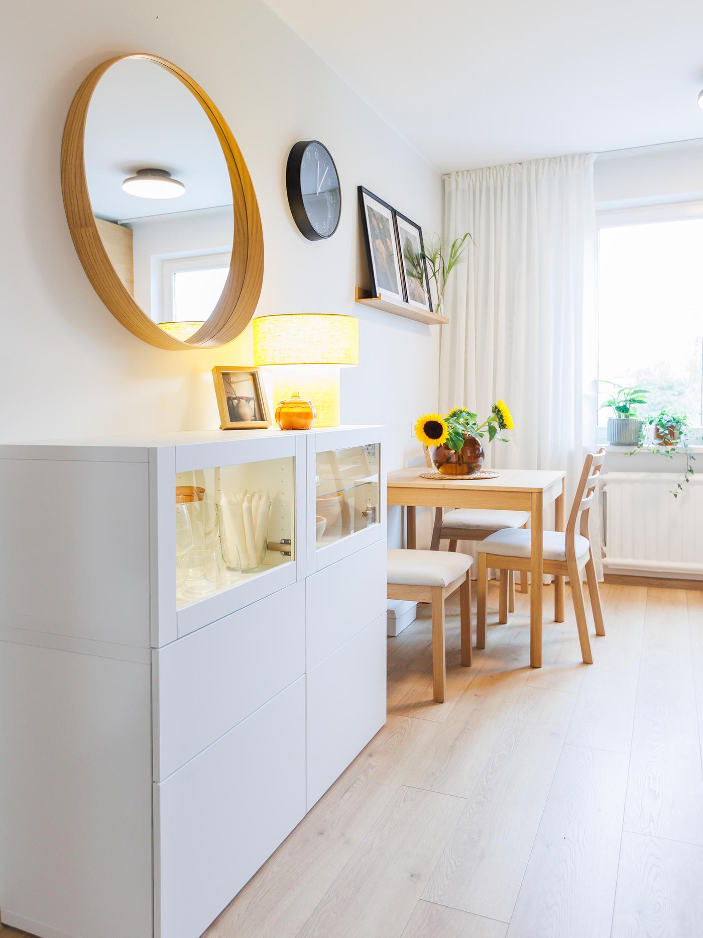 Bright dining area with a small wooden table, two chairs, a white sideboard and a round mirror on the wall.