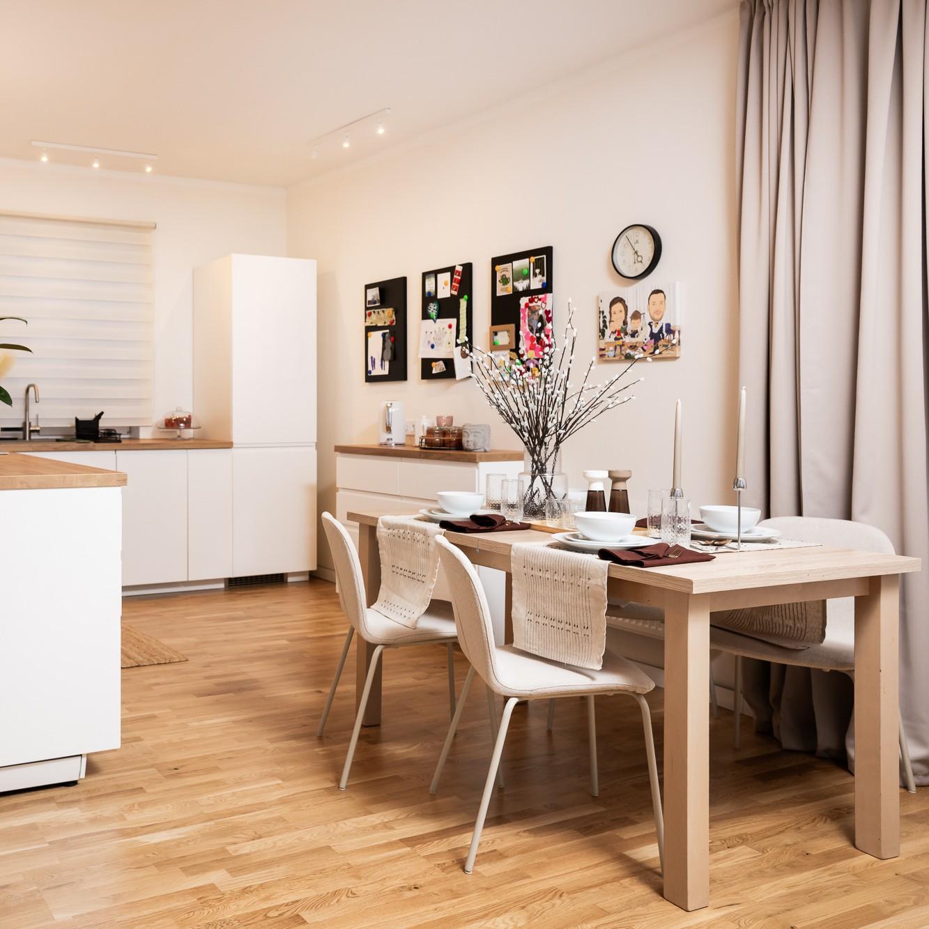 Bright dining area with a wooden table and light-coloured chairs, a white display cabinet and kitchen in the background; a tidy and harmonious space.