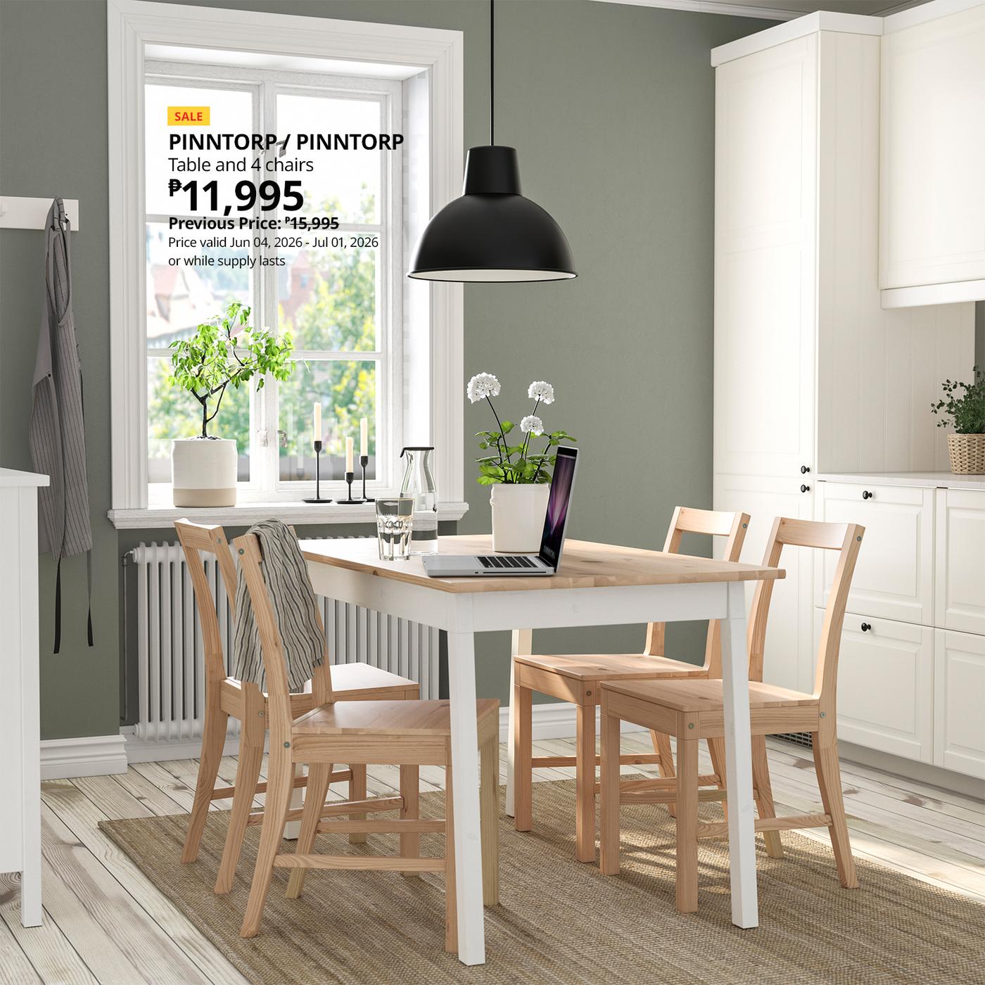 Bright dining room with a light wood table set on a beige rug, surrounded by four chairs. A laptop and glass sit on the table under a black pendant light. A window with greenery and candles is visible, along with a sale sign for the table set.