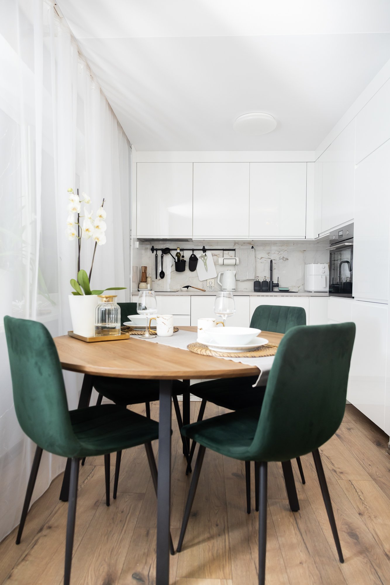 Bright kitchen with a wooden dining table and four dark green chairs; the table is set with tableware and positioned near a window with white curtains.