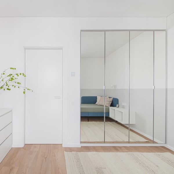 Bright, minimalist bedroom with a white door and a large wardrobe featuring mirrored sliding doors. The reflection shows a blue upholstered bench with beige throw pillows and a floating white nightstand. The light wood flooring and neutral area rug enhance the clean and modern aesthetic. A leafy branch in a vase is partially visible on the left side of the image, adding a natural touch.