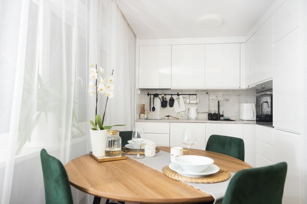 Bright, modern kitchen with white wall cabinets and built-in appliances; in the foreground there is a round wooden table set for dining with green chairs, and a white orchid by the window.