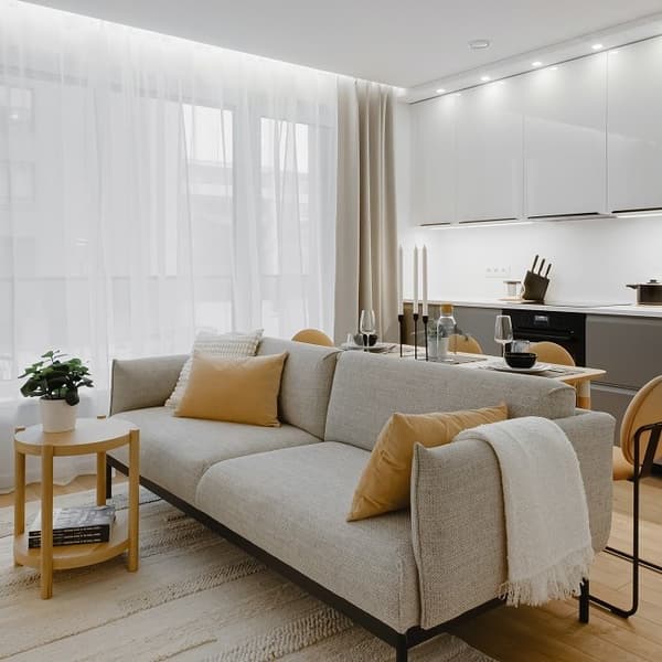 Bright, modern living room with a light grey sofa, mustard cushions, a small wooden side table, and a sleek open-plan kitchen in the background.