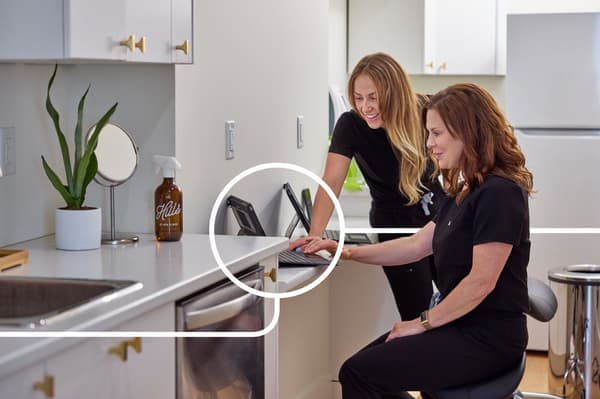 Bright, modernly designed working kitchen with IKEA furniture and decorations, two women talking in front of a tablet, a fresh green plant and a brown spray bottle as stylish accents on the white countertop.