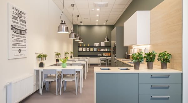 Bright office kitchen with sage green cabinets, wooden countertop, and dining area with white tables and chairs under pendant lights.