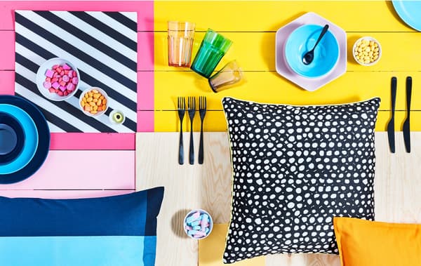 Brightly block-coloured surface with various tableware, bowls of candy and patterned cushions laid out on it.