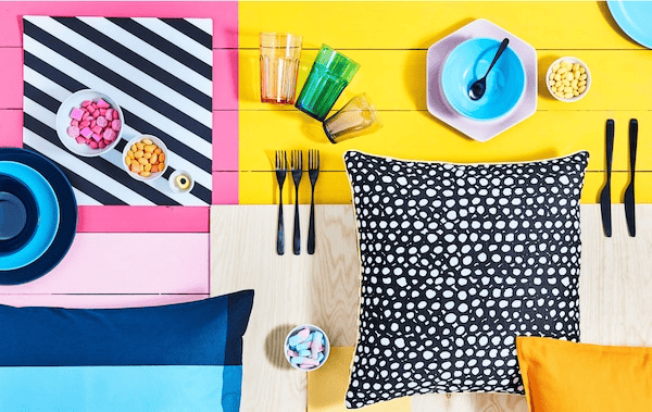 Brightly block-coloured surface with various tableware, bowls of candy and patterned cushions laid out on it.