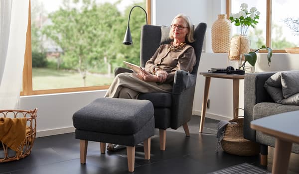Britt Monti așezată într-un fotoliu OMTÄNKSAM în colțul însorit al unei camere cu ferestre mari. Pe fundal se vede o grădină.