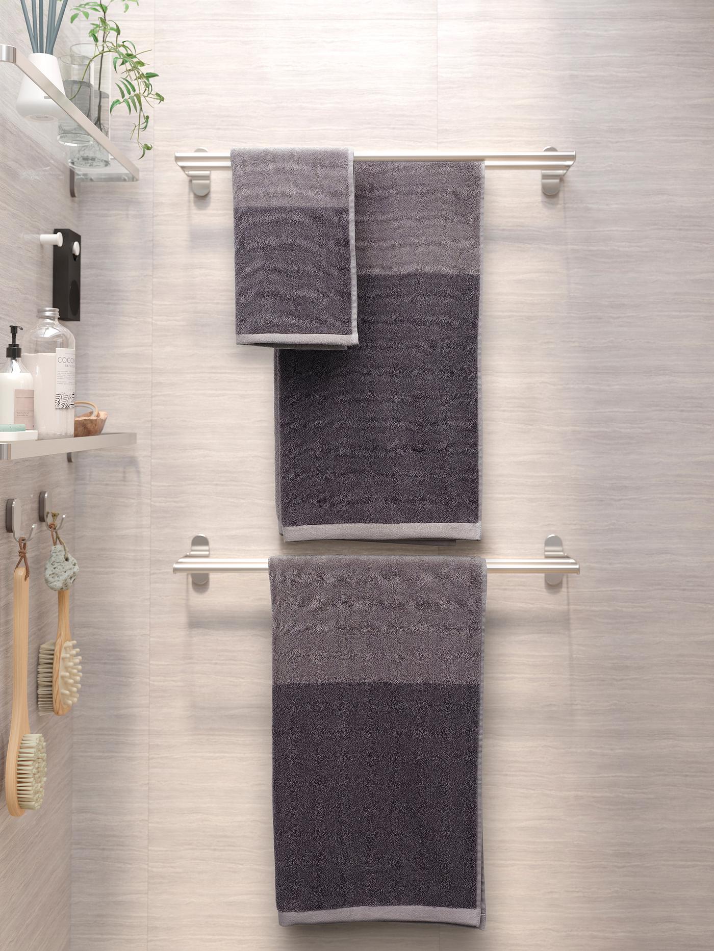 שלוש מגבות אפורות תלויות על שני מתלי מגבות BROGRUND. על הקיר משמאל מדפי זכוכית עם מוצרי רחצה.