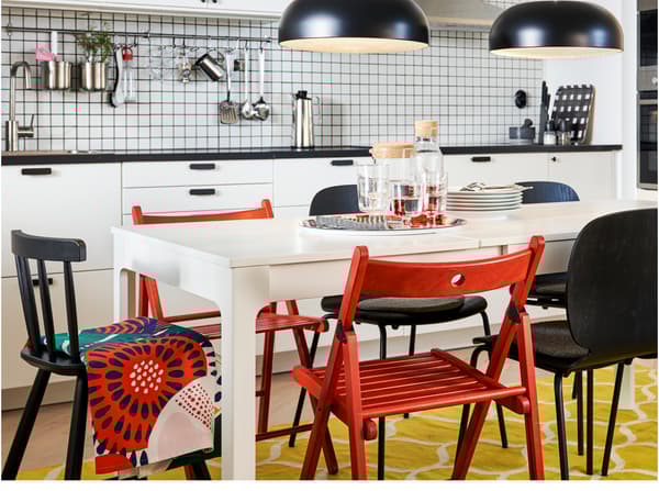 Bucătărie cu masă extensibilă desfăcută, cu scaune albe, roșii și negre, un covor galben/alb și două lustre negre.