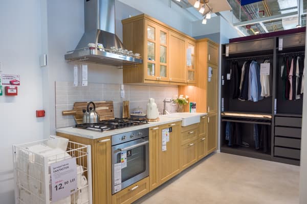 Bucătărie modernă cu dulapuri din lemn deschis, chiuvetă albă, aragaz și cuptor inox; în stânga un coș de rufe cu preț, în dreapta un dulap deschis cu haine și rafturi.