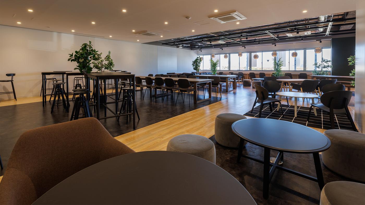 ブラックのテーブルの周りにスツールが並んでいる。奥にはハイテーブルとハイチェアが置いてある。右手奥には窓が見える。