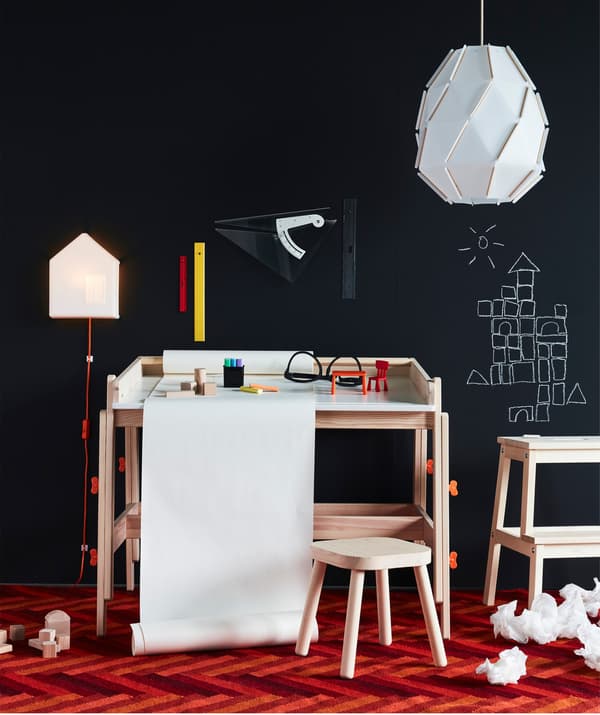 Bureau pour enfant IKEA FLISAT en bois avec support pour rouleau de papier à dessin intégré, devant un mur noir.