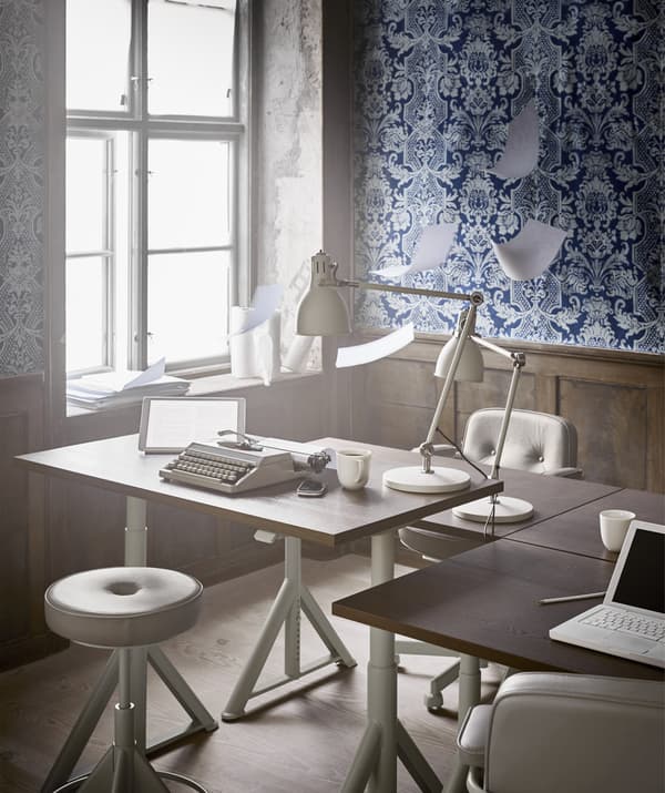 Bureau réglable en hauteur et chaise pivotante à côté d'une fenêtre dans un bureau décoré d'un papier peint bleu à motifs.
