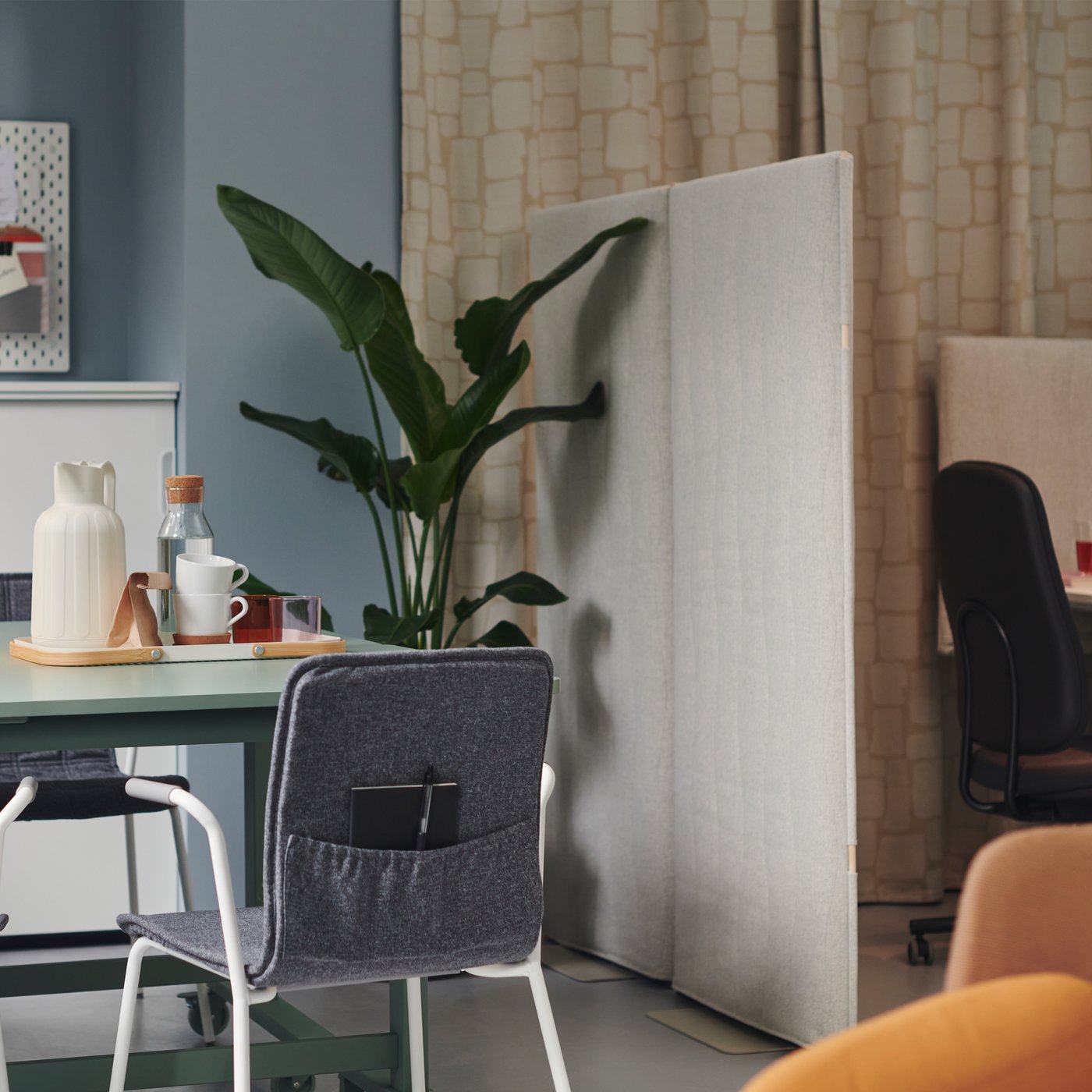 ブルーの壁の小さなオフィス。ベージュのカーテンが掛けてあり、ポケットの付いたグレーのLÄKTARE/レクタレ チェア 、コーナーに大きなグリーンの植物がある。 