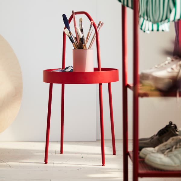 BURVIK side table in red where painting brushes are stored. BURVIK is standing next to a white wall in a bedroom.