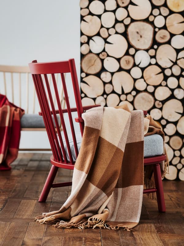 Butaca de madera roja con cojín blanco y manta de lana de líneas en tonos tierra. La pared imita a unos leños de madera apilados.