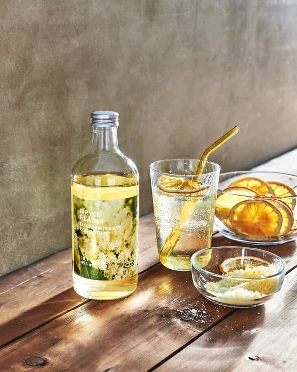 Butelka żoltego syropu z IKEA stojąca na stole, za nia szklanka z napojem i miseczka z pomarańczami