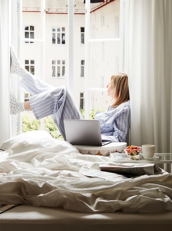 เตียงที่ยังไม่ได้ทำอยู่ถัดจากหน้าต่างที่เปิดอยู่และมีแสงแดดส่อง ผู้หญิงนั่งอยู่บนขอบหน้าต่าง มีคอมพิวเตอร์อยู่บนที่วางแล็ปท็อป BYLLAN/บิลลอน ที่อยู่ข้าง ๆ