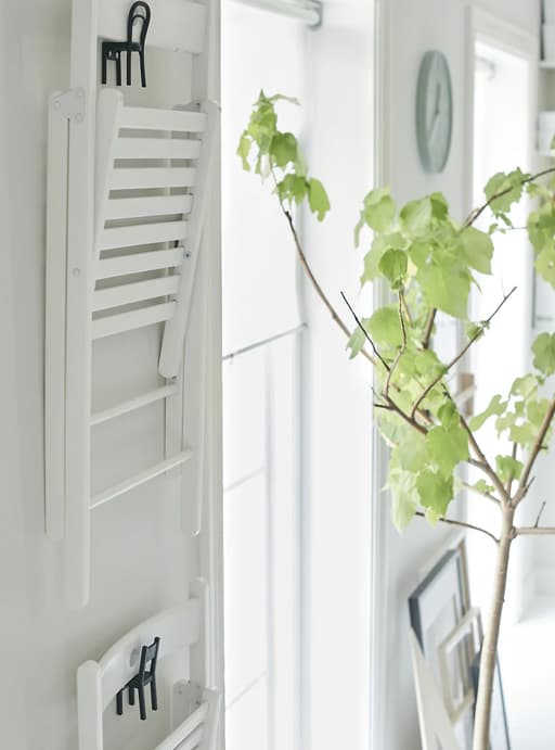 Cadires plegables blanques que pengen de ganxos en forma de minicadires a una paret blanca al costat d’una planta.