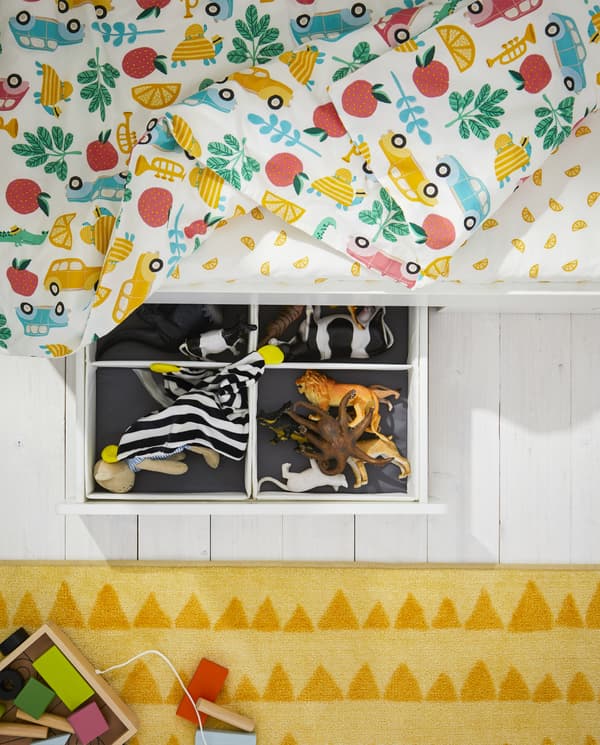 Cadre de lit avec un tiroir ouvert montrant des jouets à l’intérieur, un tapis jaune, une housse de couette à motif multicolore et un drap de lit blanc/jaune.