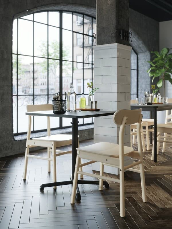 Café mit großer Fensterfront, dunklen Tischen und hellen Stühlen aus Holz.