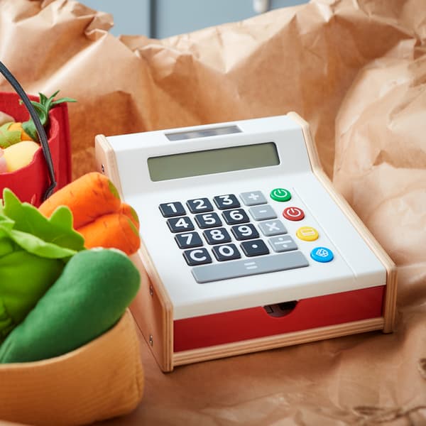 Caisse enregistreuse jouet DUKTIG et quelques légumes en peluche DUKTIG posés sur une feuille de papier kraft.