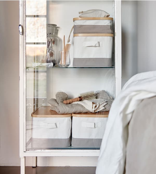 Caixas de almacenamento brancas e grises RABBLA, de bambú e produtos téxtiles, dentro dun armario branco e vintage con porta de vidro.