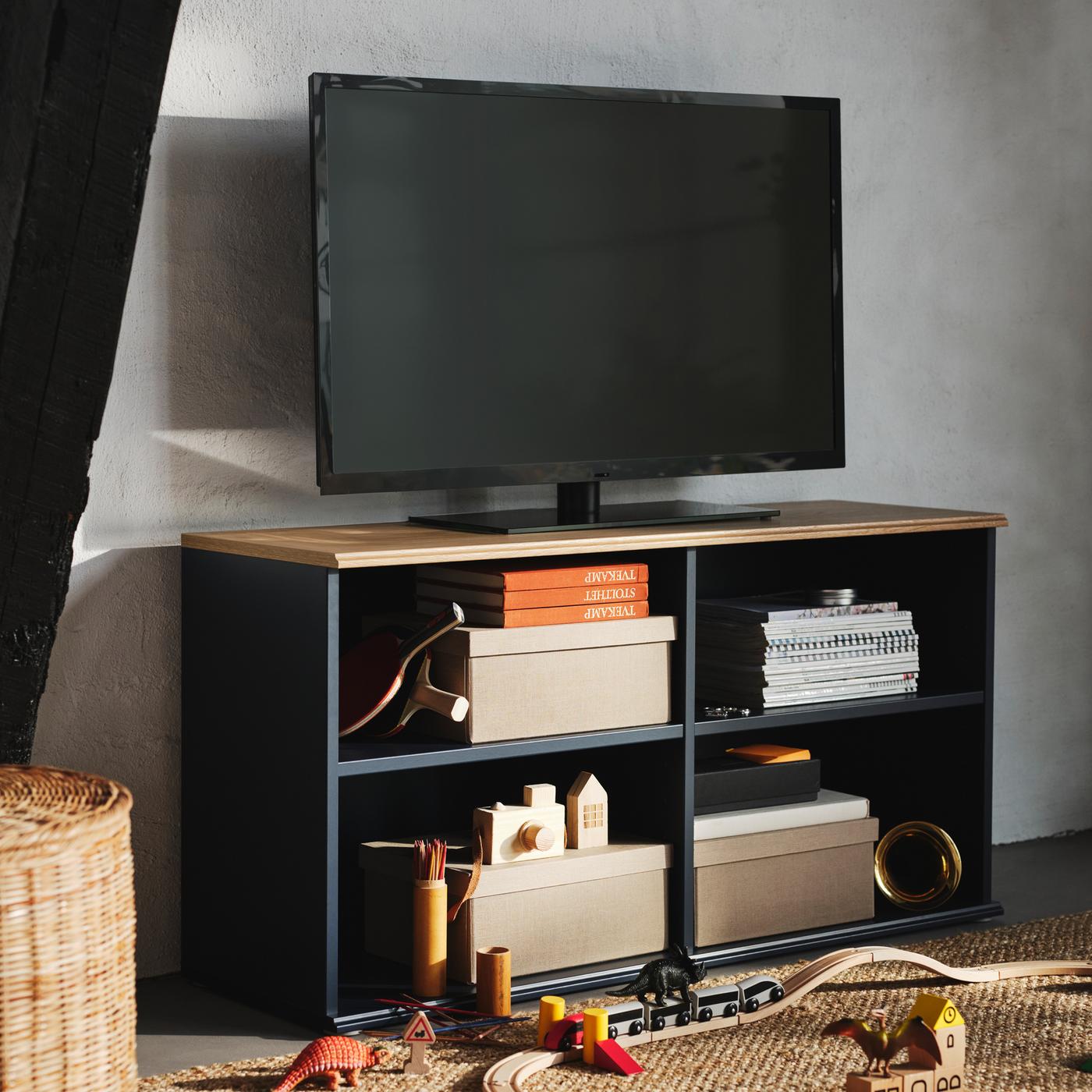 Caixas, libros e xoguetes nos estantes abertos dun moble de TV SKRUVBY de cor negra-azul, cun televisor negro enriba.