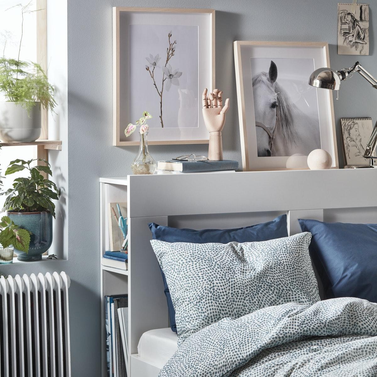 Cama BRIMNES blanca con un respaldo que tiene almacenaje incorporado. Además, tiene cuadros y decoración en la parte superior.