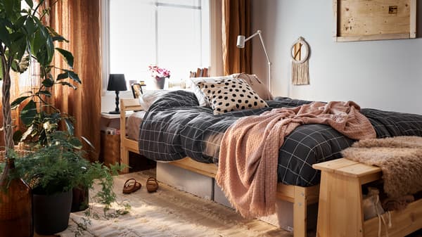 Cama com estrutura de cama em pinho com roupa de cama em preto e branco/xadrez, caixas de arrumação em branco/cinzento e um tapete por baixo.