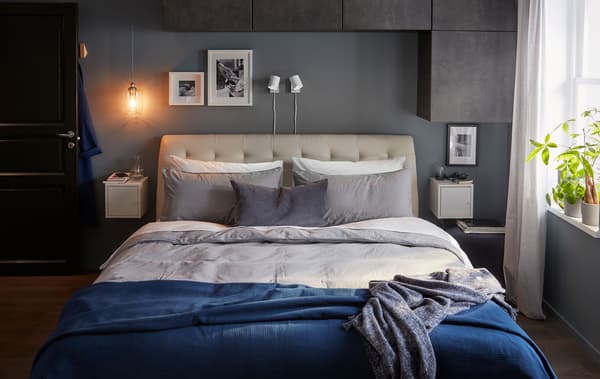 Cama dobre nun dormitorio en tons grises e azuis. Armarios en parede sobre a cama e xunto a ela; plantas na ventá.