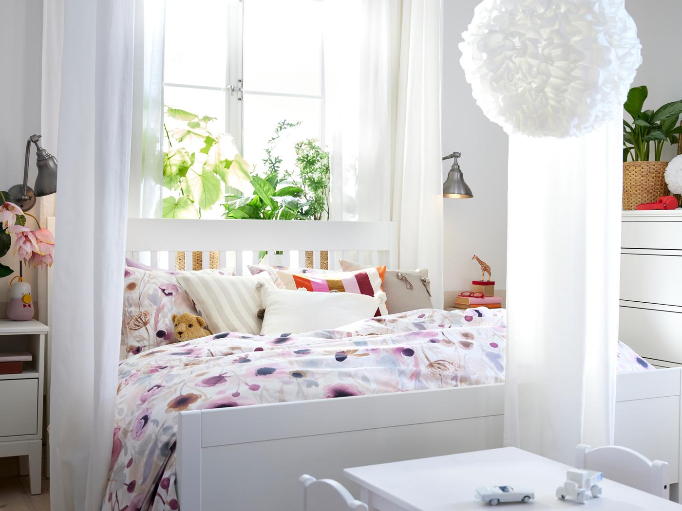 Cama IDANÄS em branco com roupa de cama LÖNNHÖSTMAL em frente a janela. Há uma mesa para criança aos pés da cama.