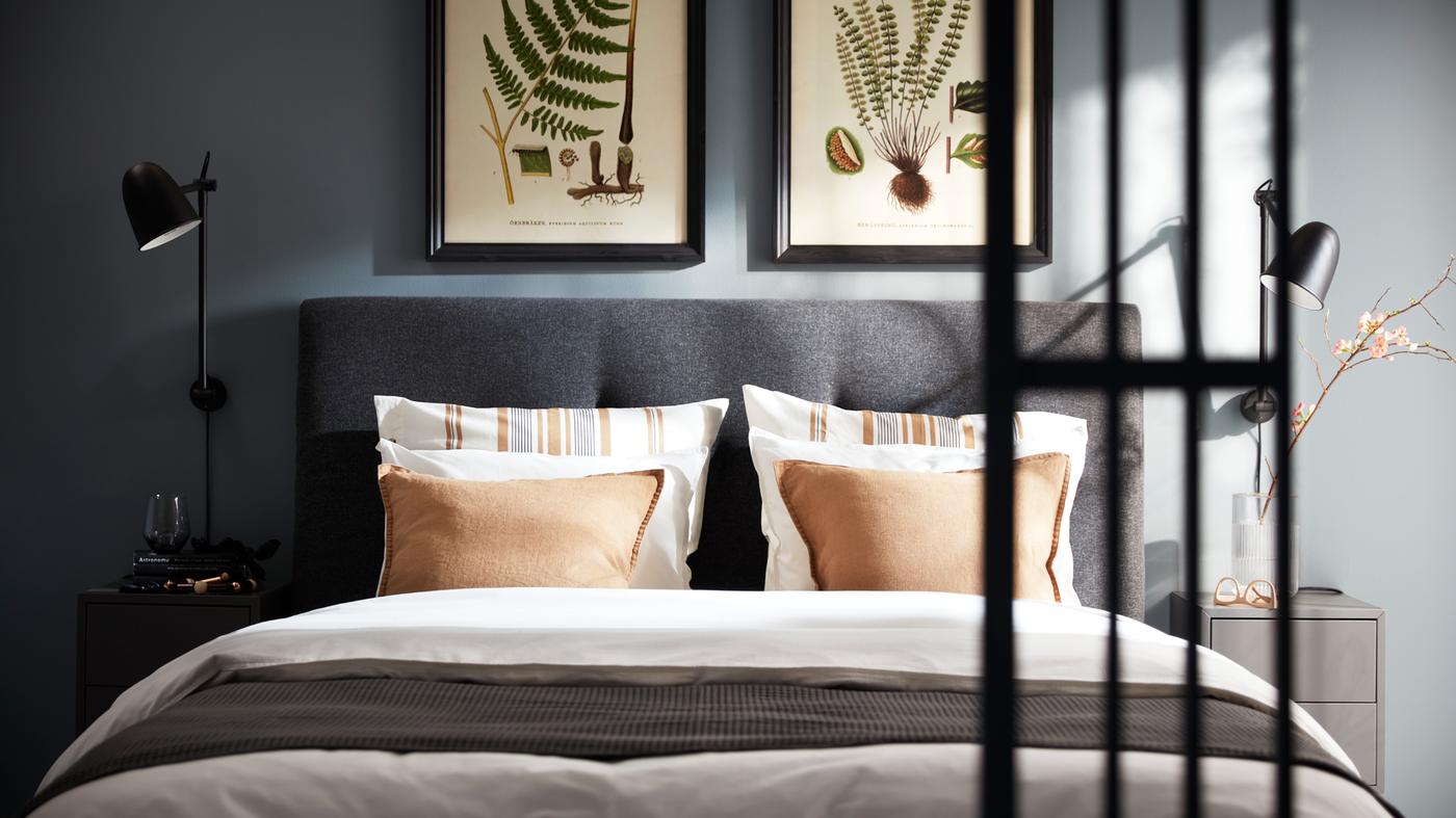 Cama IDANÄS em cinzento escuro com roupa de cama ÄNGSLILJA em branco, colcha VÅRELD em cinzento escuro e candeeiros de parede SKURUP em preto em cada um dos lados.