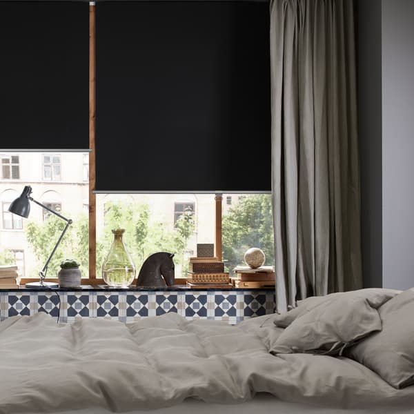 Cama no centro de quarto, janelas com cortinados em cinzento e estores de correr opacos TRETUR descidos até meio.