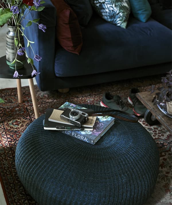 Camera and books on a large round blue pouffe, with blue sofa in the background.