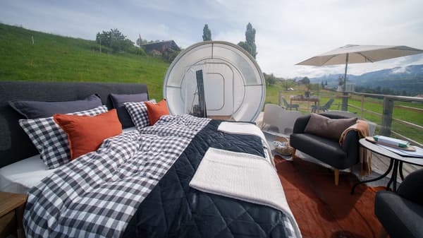 Camera da letto accogliente con una trapunta a quadri bianchi e neri, cuscini arancioni e vista sulle colline verdi e sulle montagne attraverso pareti trasparenti con cuscini d'aria.