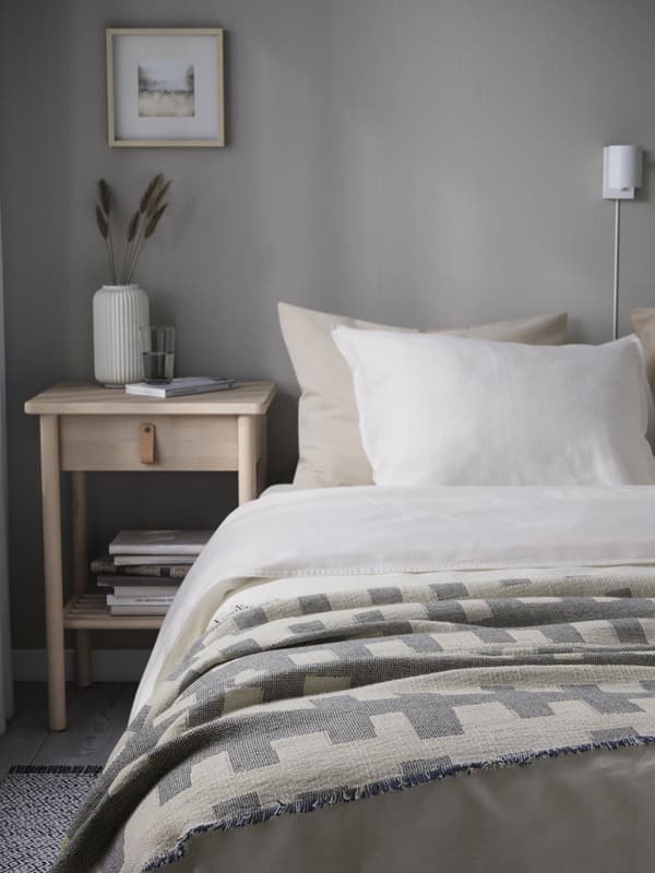 Camera da letto con biancheria sui toni chiari del beige, bianco e grigio. Accanto, un comodino con libri e un vaso.