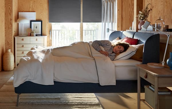 Camera da letto con una donna addormentata in un letto TUFJORD blu mentre la luce del sole filtra dalla finestra alle sue spalle.