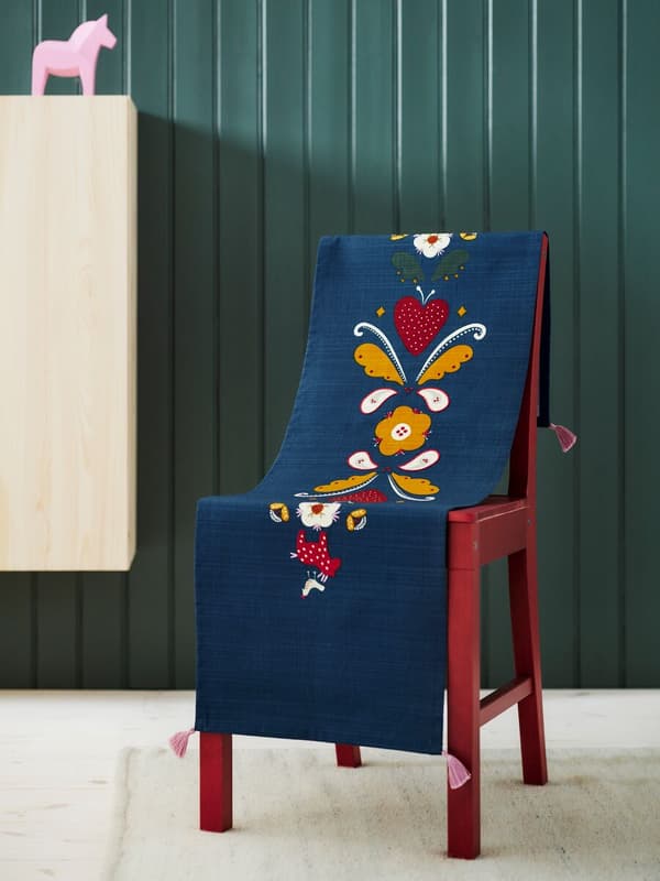 Camino de mesa azul con motivos coloridos sobre una silla de madera roja. Pared verde de madera y un muble de madera cruda con un adorno en forma de caballo de color rosa.